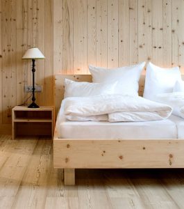 Minimalistisches Schlafzimmer mit hellen Holzböden und -wänden, einem einfachen Holzbett mit weißem Bettzeug und Kissen, einem kleinen hölzernen Nachttisch und einer schwarzen Lampe mit weißem Lampenschirm.