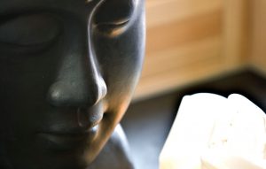 Nahaufnahme einer glatten, dunklen Statue eines heiteren Gesichts, möglicherweise Buddha, mit weichem Licht und unscharfen weißen und beigen Objekten im Hintergrund.