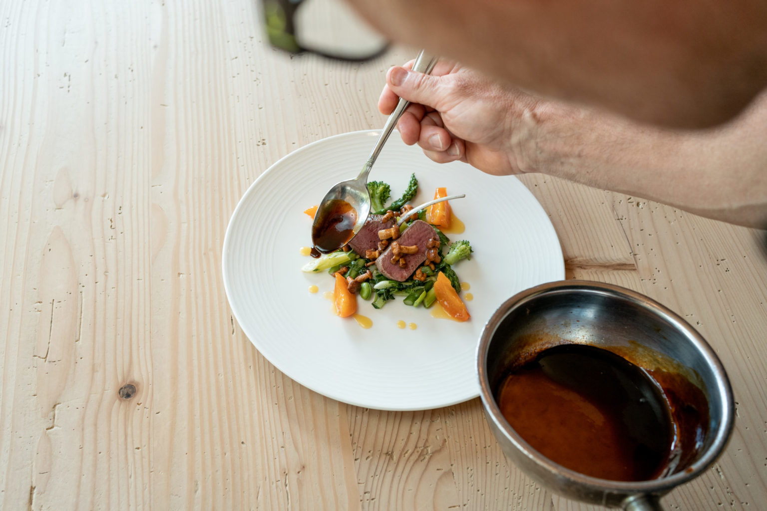 Eine Person löffelt vorsichtig Soße auf eine Platte mit Fleisch- und Gemüsescheiben auf einem weißen Teller, während ein Soßentopf in der Nähe auf einem Holztisch steht.