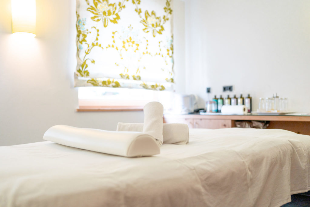 Ein Massagetisch mit einem weißen Laken und ordentlich aufgerollten weißen Handtüchern in einem hellen, aufgeräumten Raum mit geblümten Vorhängen und Flaschen auf einem Holztisch im Hintergrund.