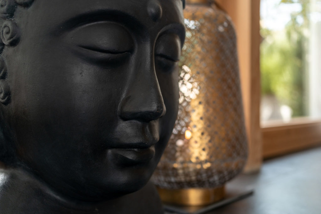 Nahaufnahme des Gesichts einer dunklen, ruhigen Buddha-Statue mit geschlossenen Augen, neben einer geflochtenen Laterne mit weichem, warmem Licht und einem Fenster mit unscharfem Grün im Hintergrund.
