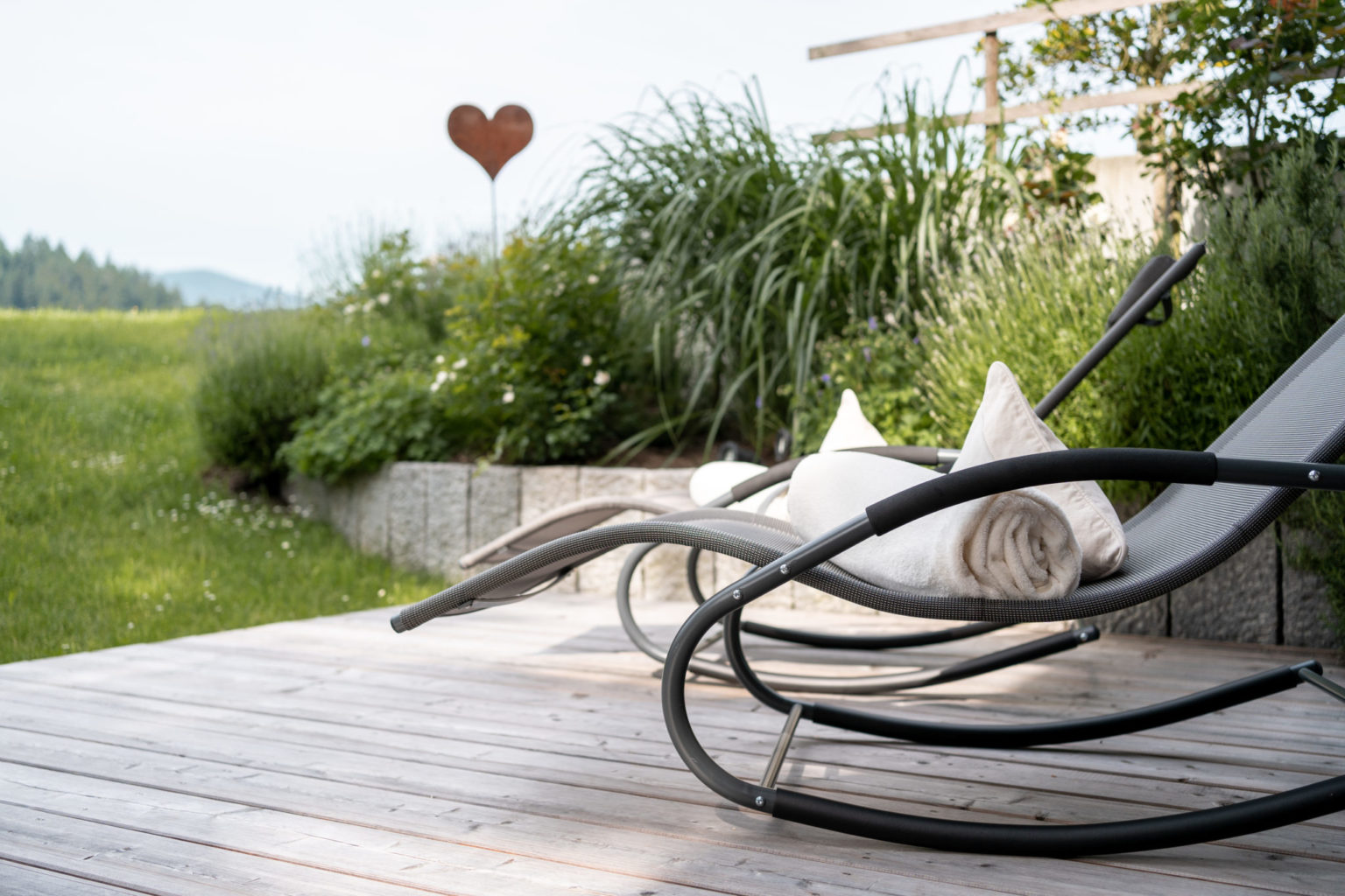Zwei moderne Schaukelstühle mit gerollten Handtüchern stehen auf einer Holzterrasse mit Blick auf einen üppigen Garten und eine grüne Wiese, mit einer herzförmigen Skulptur im Hintergrund.