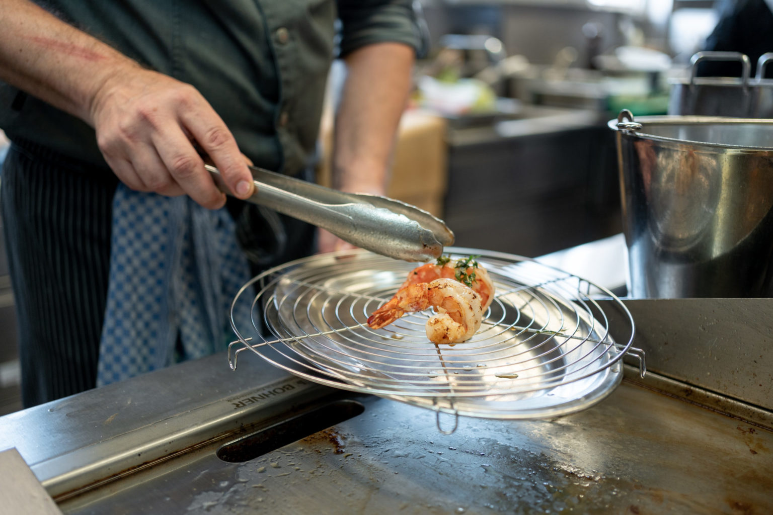 Ein Koch benutzt eine Zange, um gekochte Garnelen auf ein rundes Drahtgestell über einem Grill in einer professionellen Küche zu legen.