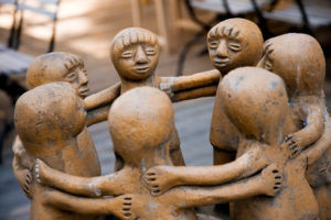 Eine Nahaufnahme einer runden Tonskulptur, die sechs menschliche Figuren darstellt, die im Kreis stehen und sich gegenseitig die Arme um die Schultern legen, um Einheit und Zusammengehörigkeit zu symbolisieren.